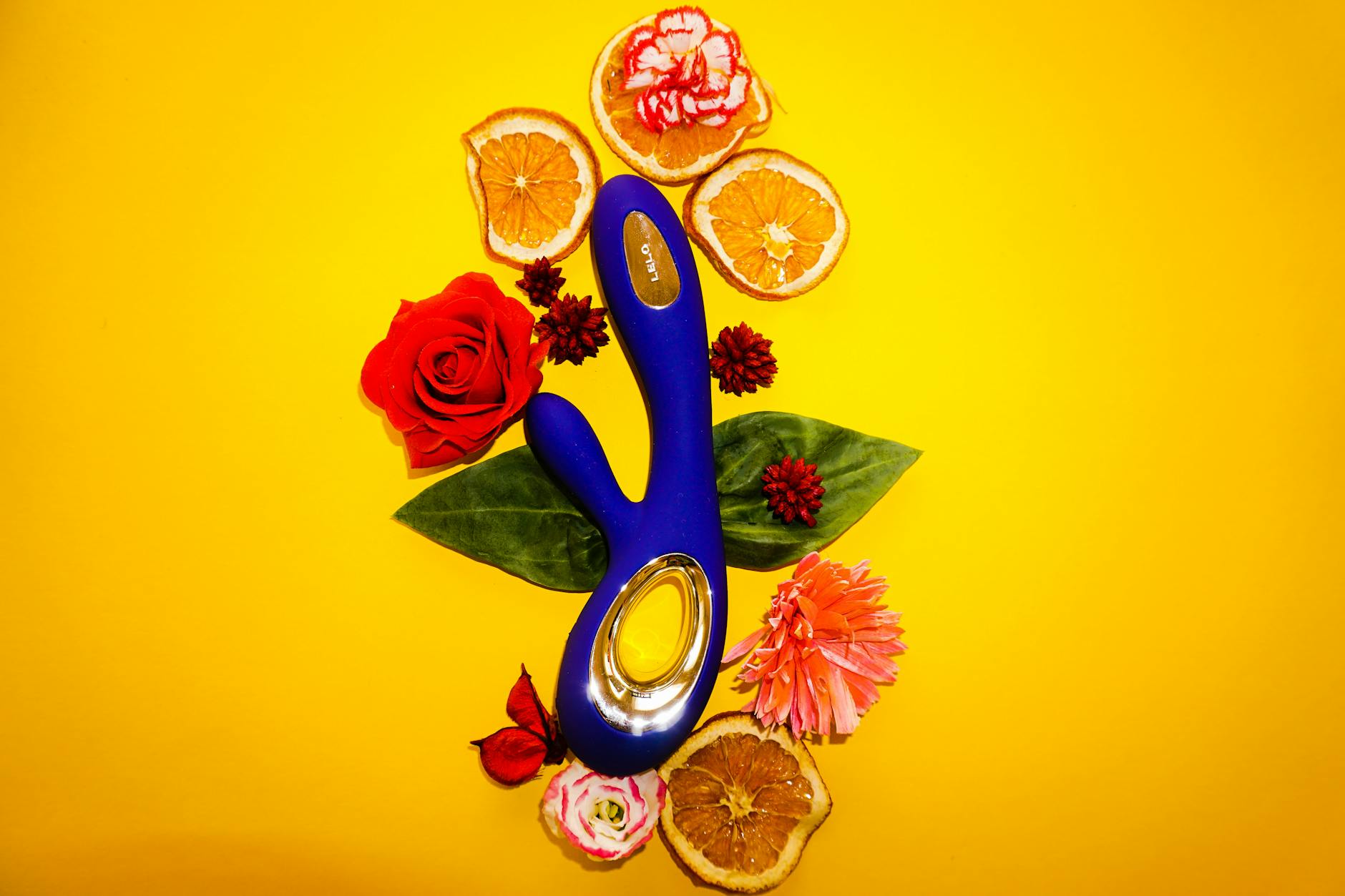 A blue vibrator surrounded by fresh flowers and citrus slices on a bright yellow background